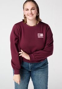 Bordeauxrode trui met geribbelde textuur en opgeblazen mouwen, met een klein roze grafisch ontwerp aan de voorkant. Draagt blauw spijkerbroek.