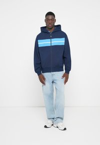 Mørkeblå lynlås hoodie med en blå vandret stribe, der har hætte og ribbede manchetter. Sære med lyseblå jeans og grå sneakers.