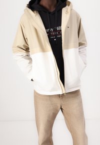 Lekka kurtka w kolorach beżowym, kremowym i białym z przednim zapięciem na suwak, kapturem i kontrastującym wzorem kolorystycznym. Noszona na czarnej bluzie z kapturem.