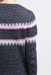 Strickpullover mit marineblauem Grund und geometrischen Mustern in Weiß, Pink und Hellblau auf dem oberen Rücken und den Ärmeln, getragen von einer Person.
