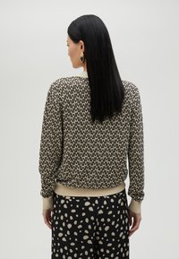 Džemper s uzorkom na bež pozadini i crnim apstraktnim dizajnom, rebrastim manžetama i strukom; uparen s crnim hlačama s točkicama.