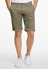 Pantaloni corti di cotone verde oliva con risvolti, tasche frontali e chiusura con bottone, abbinati a sneaker bianche.