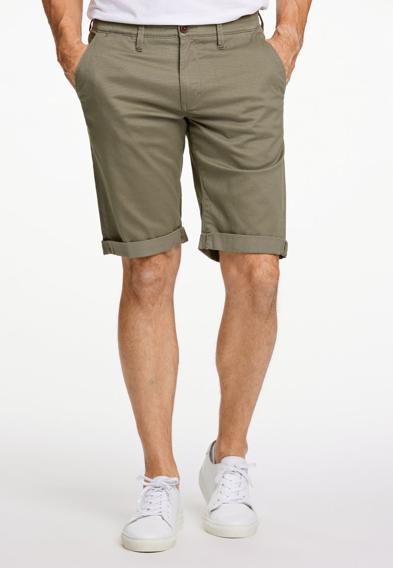 Pantaloni corti di cotone verde oliva con risvolti, tasche frontali e chiusura con bottone, abbinati a sneaker bianche.