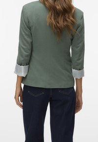 Femme portant un blazer vert sauge avec une doublure de poignet à rayures et un jean en denim bleu foncé, vue de dos sur un fond blanc.