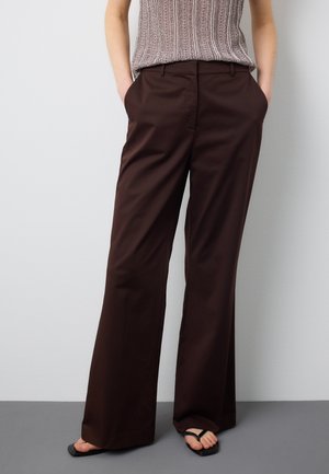 Frau trägt dunkelbraune weite Hosen, beige ärmelloses Strickoberteil, schwarze Sandalen mit quadratischer Spitze, steht mit den Händen in den Taschen.