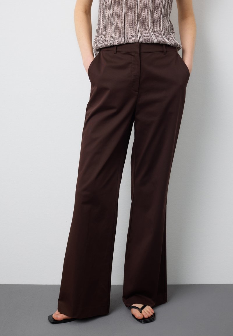 Frau trägt dunkelbraune weite Hosen, beige ärmelloses Strickoberteil, schwarze Sandalen mit quadratischer Spitze, steht mit den Händen in den Taschen.