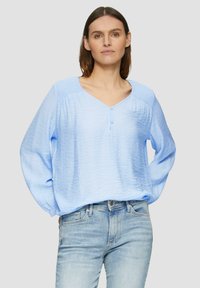 Blouse bleu clair avec un col en V et des détails de boutons, fabriquée en tissu texturé et à manches longues, assortie à un jean bleu clair.