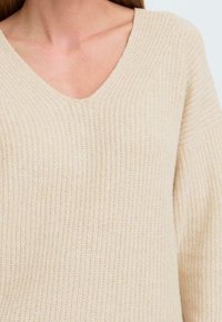Pull en tricot beige avec un col en V et une texture côtelée. Le tissu semble doux et léger avec une coupe décontractée.