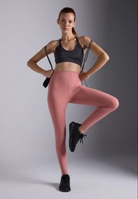 Mlada žena u crnom sportskom grudnjaku i ružičastim leggingsem drži konopac za preskakanje, stojeći na jednoj nozi u izbalansiranoj fitness pozi.