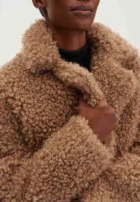 Cappotto texture riccio marrone con colletto alto, realizzato in un morbido tessuto peloso. Una mano sta stringendo il cappotto vicino al colletto.