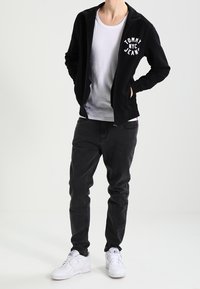 Sudadera negra con cremallera y el logo "TOMMY NYC JEANS", usada sobre una camiseta blanca. Combinada con vaqueros oscuros y zapatillas blancas. Fondo neutro.