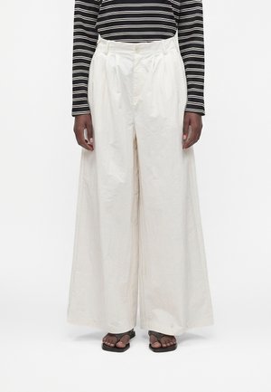 Persona che indossa pantaloni bianchi a vita alta e gamba larga, top a maniche lunghe a righe bianco e nero e sandali neri con punta aperta, in piedi su sfondo bianco.