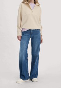 Sudadera de forro polar color crema con media cremallera, combinada con una camisa lila clara, pantalones vaqueros azules de pierna ancha y zapatillas blancas.