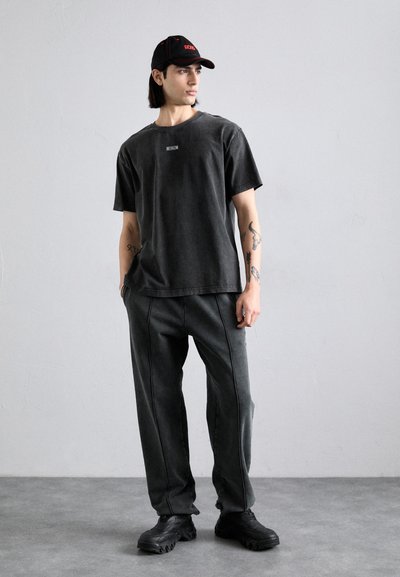 T-shirt noir oversized avec une petite étiquette, pantalon gris foncé avec des coutures latérales, et baskets noires chunky, porté avec une casquette noire avec des accents rouges.