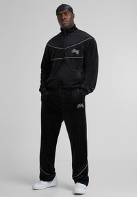 Trening din velur negru cu baretă albă, jachetă cu fermoar și pantaloni cu talie elastică. Prezintă logo pe piept și pe șold; asortat cu adidași albi.