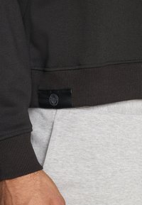Sweatshirt cinza escuro com punhos e barra canelados, apresentando um pequeno rótulo preto na parte inferior. Tecido cinza das calças visível abaixo.