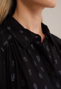 Zwart overhemd met kraag, voorzien van een gestructureerde stof en zilverkleurig gestippeld patroon. Knopen aan de voorkant en een lichte plooivorming bij de schouders.