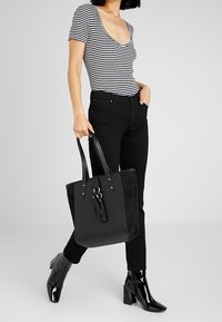 Czarna torba na ramię o gładkim wykończeniu i zamszowych wstawkach, z długimi czarnymi uchwytami i akcentem w postaci frędzla. Noszona z paskowanym topem i czarnymi dżinsami.