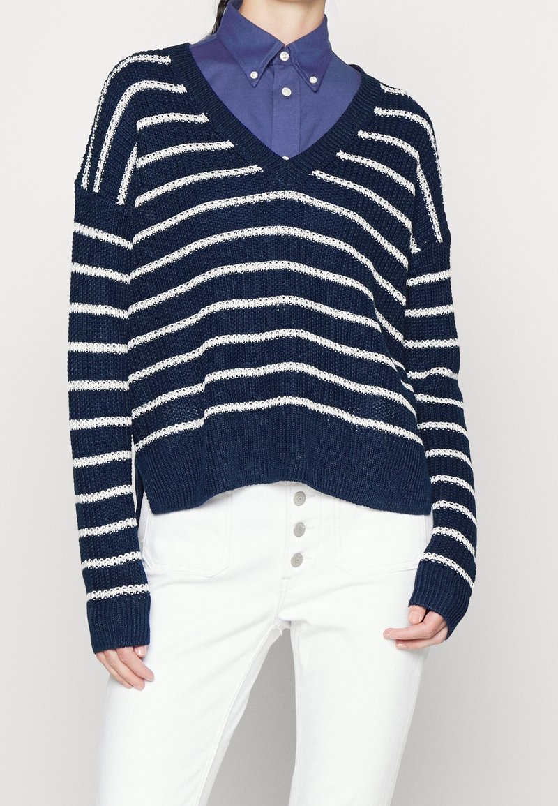 Personne portant un pull à col en V rayé bleu marine et blanc sur une chemise bleue boutonnée, associé à un pantalon blanc taille haute.