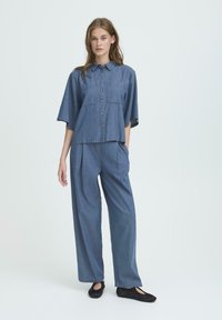 Denimhemd mit kurzen Ärmeln und Knopfverschluss, kombiniert mit weit geschnittenen Denim-Hosen, beide in einem mittelblauen Farbton und mit Taschen.