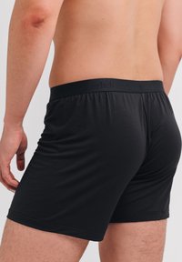Schwarze Boxershorts mit glatter Textur, einem elastischen Bund mit eingeprägtem Logo und einer figurbetonten Passform, die die Körperform betont.