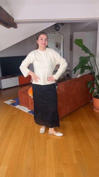 Jeune femme debout dans le salon, les mains sur les hanches, portant un pull blanc, une jupe noire, des chaussons blancs, près d'un canapé orange et d'une grande plante verte.