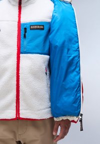 Giacca in pile con corpo bianco, maniche blu e dettagli rossi. Caratterizzata da una tasca sul petto e polsini elasticizzati. Chiusura con zip e logo del marchio visibile.