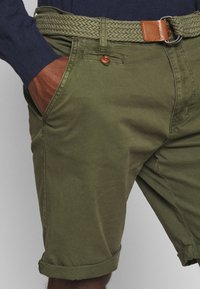Shorts en coton vert olive avec une ceinture tressée, dotés d'une finition en cuir marron, de poches latérales et de revers roulés.
