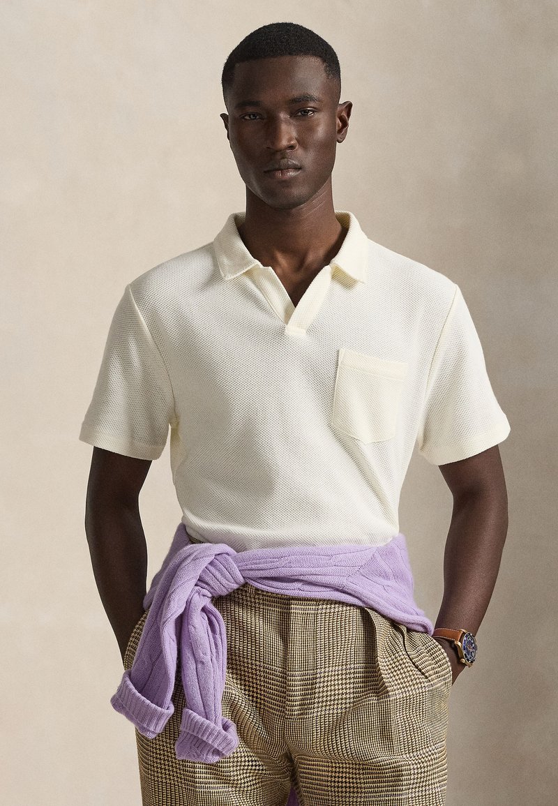 Άνδρας που φοράει εκρού πόλο με κοντά μανίκια, μπεζ καρό παντελόνι, λιλά πουλόβερ δεμένο γύρω από τη μέση του και ρολόι χειρός, στέκεται μπροστά σε ουδέτερο φόντο.