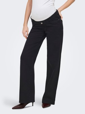 Personne enceinte portant un pantalon noir à jambes larges et un haut blanc cintré, debout, une main sur la hanche, portant des talons pointus marron.