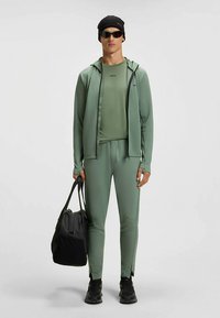 Sportlik komplekt tuhmunud rohelisest kangast, mis koosneb lukuga kapuutsiga jopest, kehaümbrilisest särgist ja kitsenevatest pükstest. Aksessuaarideks must kott ja päikeseprillid.