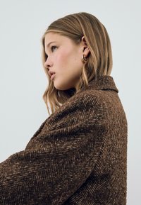 Manteau en laine texturée marron avec un motif en chevron, col large et épaules tombantes, présentant une vue de profil d'une personne le portant.