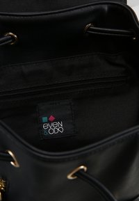 Intérieur d'un sac seau en cuir noir comprenant une poche zippée et une étiquette en tissu affichant le nom de la marque "even & odd" avec des accents colorés.