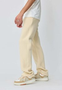 Pantalons de survêtement beige, coupe décontractée avec une texture lisse, dotés de poches latérales et d'une jambe droite, associés à des baskets beiges et blanches.