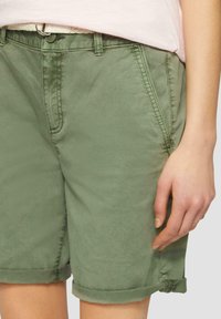 Shorts en coton verts avec ourlets roulés, poches avant et détails de couture visibles. Le matériau semble lisse avec une légère brillance.