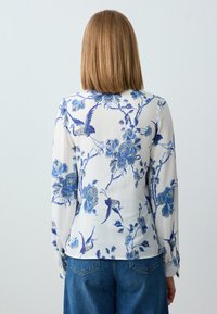 Frau mit schulterlangem, glattem Haar, die eine weiße Bluse mit blauen Blumen- und Vogelmotiven trägt, kombiniert mit blauen Jeans, von hinten betrachtet.