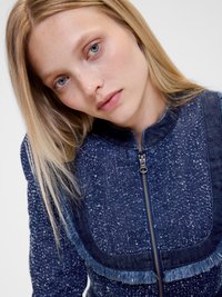 Giacca di jeans blu scuro con una texture a macchie. Presenta una chiusura con zip, dettagli in frange lungo il colletto e un design strutturato.