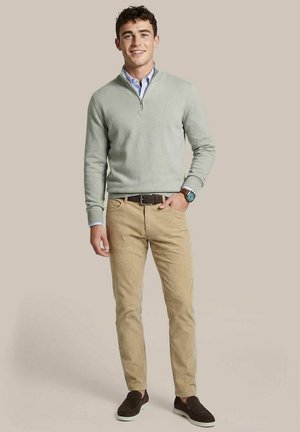 Young man smiling, wearing light gray half-zip sweater, blue collared shirt, beige pants, dark slip-on shoes, and wristwatch, standing on neutral background.