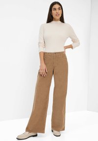 Haut long à manches longues beige côtelé associé à un pantalon en velours côtelé marron à jambe large. Des chaussures marron complètent la tenue, avec un détail de bouton sur le pantalon.