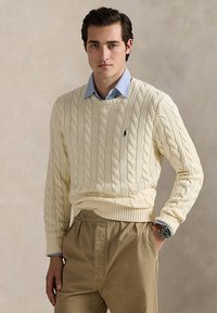 Hombre con suéter de punto trenzado color crema sobre camisa azul con cuello, pantalones caqui, mano en el bolsillo y reloj en la muñeca izquierda.