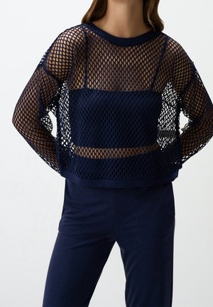 Femme portant un haut en maille bleu marine à manches longues par-dessus un crop top bustier assorti et un pantalon bleu marine sur un fond uni.