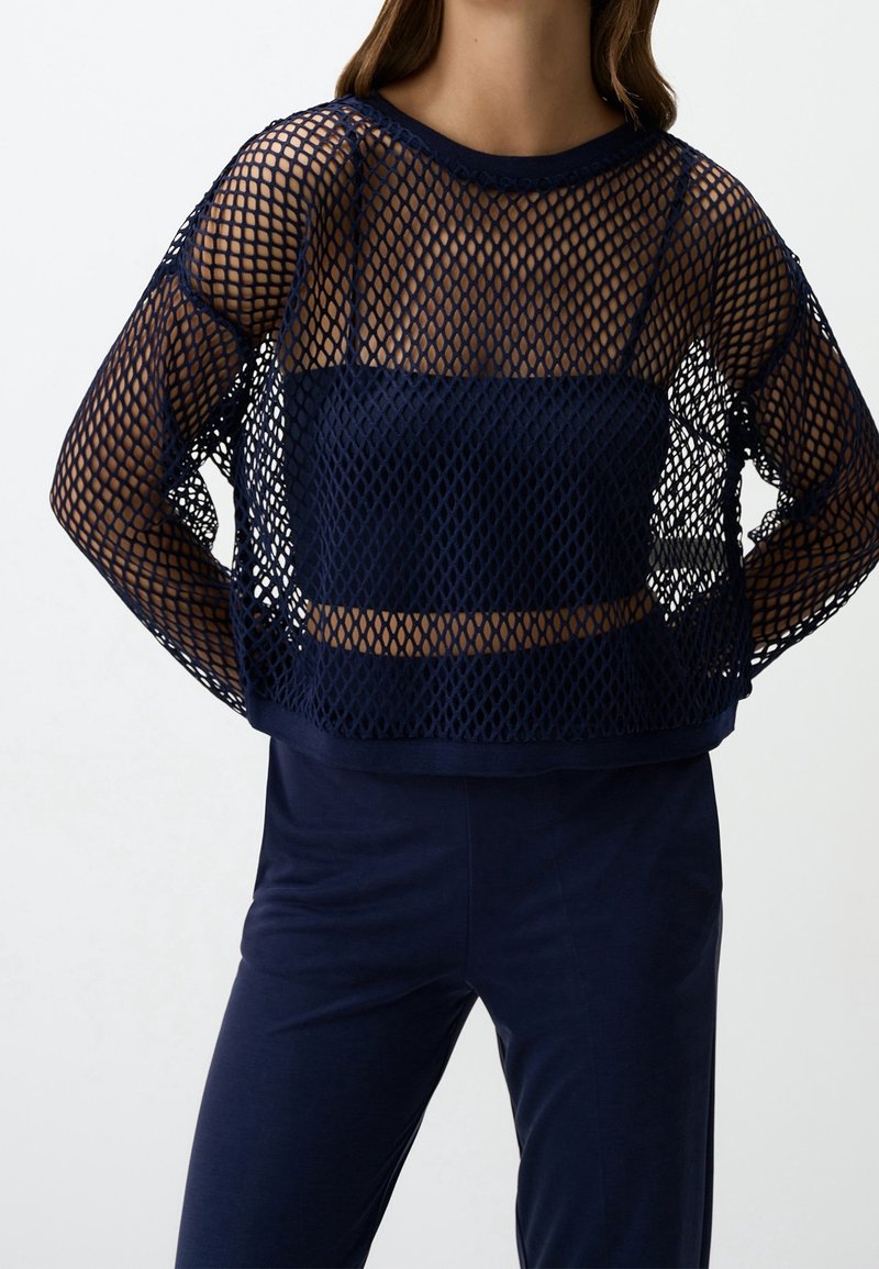 Femme portant un haut en maille bleu marine à manches longues par-dessus un crop top bustier assorti et un pantalon bleu marine sur un fond uni.