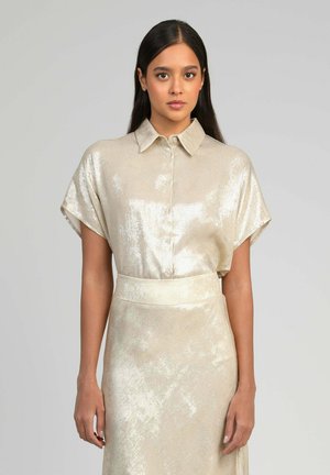 Jeune femme aux longs cheveux foncés portant un chemisier beige brillant à manches courtes avec boutons et une jupe taille haute assortie.