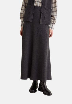 Mujer que lleva una falda larga gris oscuro, botas negras hasta el tobillo, una camisa de cuadros y un chaleco gris oscuro abotonado con bolsillos.