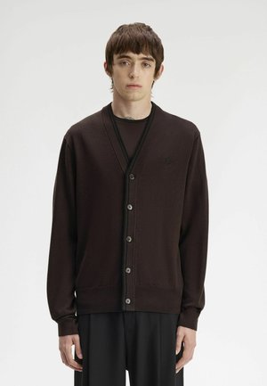 Giovane uomo con capelli castani corti che indossa un cardigan scuro abbottonato sopra una camicia nera, in piedi davanti a uno sfondo chiaro e uniforme.