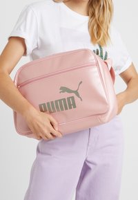 Borsa sintetica rosa con una texture liscia, caratterizzata da un grande logo Puma grigio e un compartimento con zip. Progettata con una tracolla.