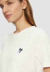 Witte T-shirt met korte mouwen, voorzien van een klein geborduurd hartontwerp in blauw en zwart op het linkse borstgedeelte.
