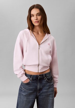 Jeune femme portant un sweat à capuche zippé rose clair et un jean bleu foncé, debout, une main dans la poche, sur un fond gris uni.