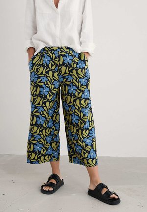 Gekürzte, gemusterte Hosen mit einem floralen Design in Blau und Grün auf einem dunklen Hintergrund, aus atmungsaktivem Stoff. Schwarze Slipper-Sandalen.