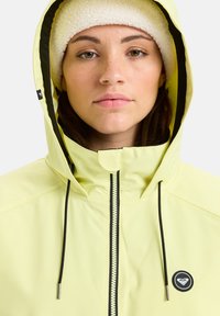 Corta-vento amarelo à prova d'água com capuz forrado de fleece, fecho éclair preto e logótipo. Apresenta ajustadores de cordão e uma textura suave.